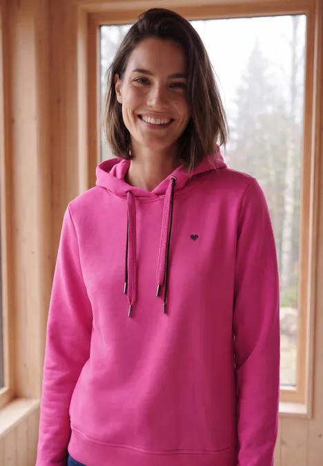 Frau in pinkem Kapuzenpulli, die lächelnd vor einem Fenster in einer Holzhütte steht.