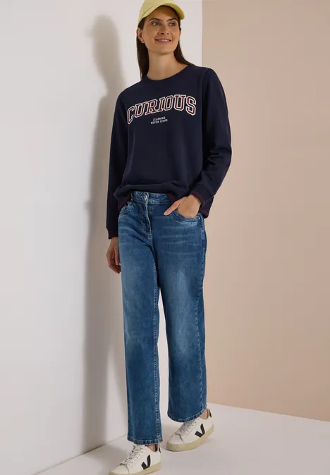 Junge Frau lehnt an einer Wand, trägt dunkelblauen Sweatshirt, gerade Jeans, weiße Sneakers und gelbe Mütze.