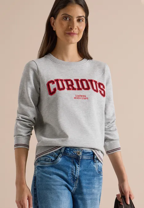 Junge Frau in grauem Sweatshirt mit rotem 'CURIOUS'-Schriftzug und blauen Jeans.