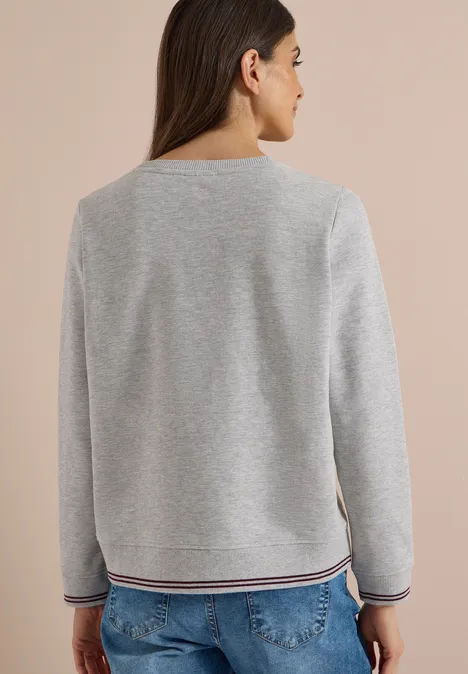 Rückenansicht einer Frau in grauem Pullover mit gestreiften Bündchen und blauer Jeans.
