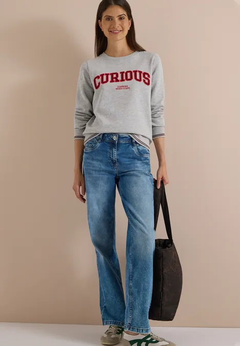 Frau in grauem 'CURIOUS'-Pullover, blauen Jeans und schwarzer Tasche, lässiger Look vor neutralem Hintergrund.