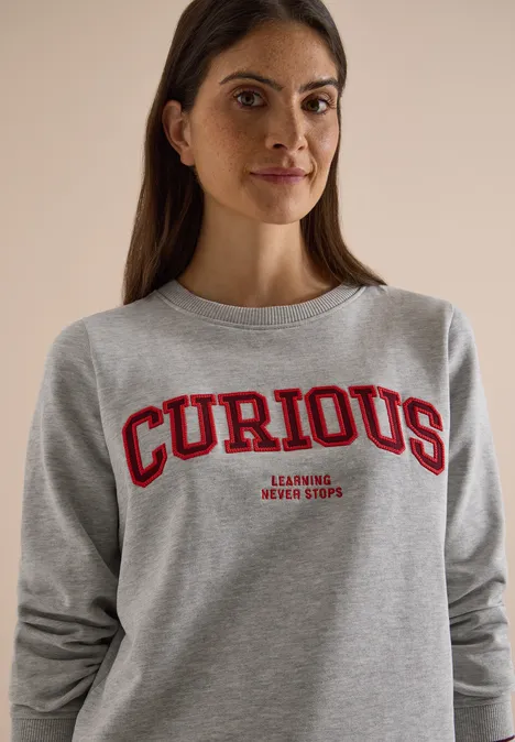Frau in grauem Pullover mit rotem 'CURIOUS'-Schriftzug und kleinem Text darunter.
