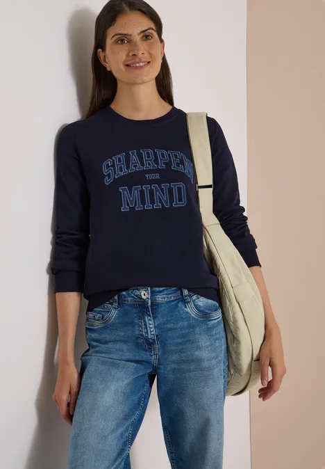 Frau in Jeans und dunkelblauem Pullover mit Aufschrift 'SHARPEN MIND', trägt eine Tasche und lächelt