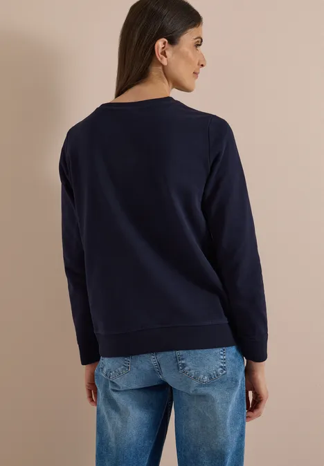 Rückansicht einer Frau in dunkelblauem Pullover und Jeans vor beigem Hintergrund.