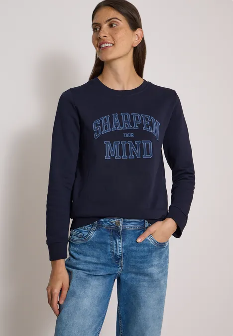 Frau trägt dunkelblauen Pullover mit 'SHARPEN MY MIND'-Schriftzug und Jeans vor neutralem Hintergrund.