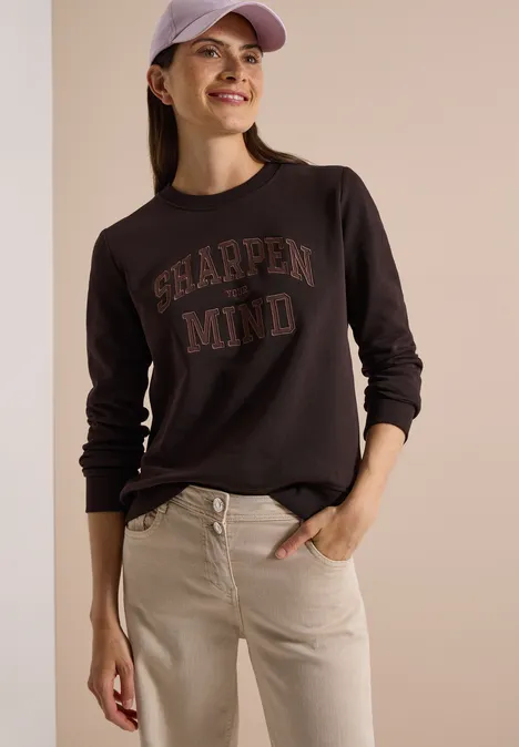 Frau in dunkelgrauem Sweatshirt mit 'SHARPEN MIND'-Aufdruck, beigen Jeans und rosa Mütze lächelt.