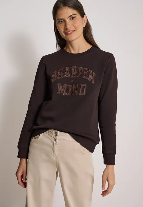 Frau in braunem Pullover mit Schriftzug 'SHARPEN MIND' und beige Hose vor hellem Hintergrund.