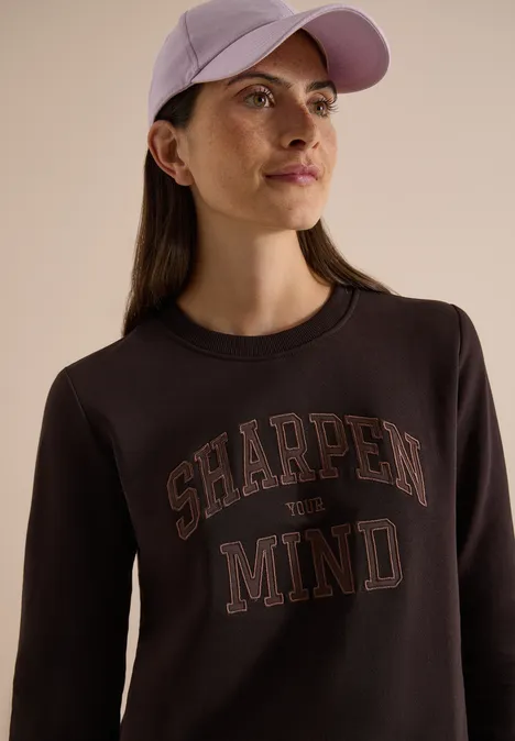 Eine Frau trägt eine helle Kappe und einen schwarzen Pullover mit der Aufschrift "SHARPEN MIND".