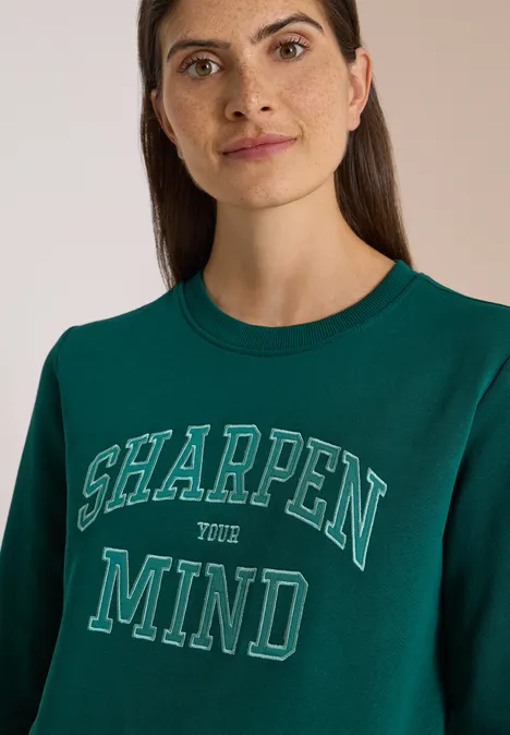 Porträt einer Frau im dunkelgrünen Sweatshirt mit der Aufschrift 'SHARPEN YOUR MIND'.