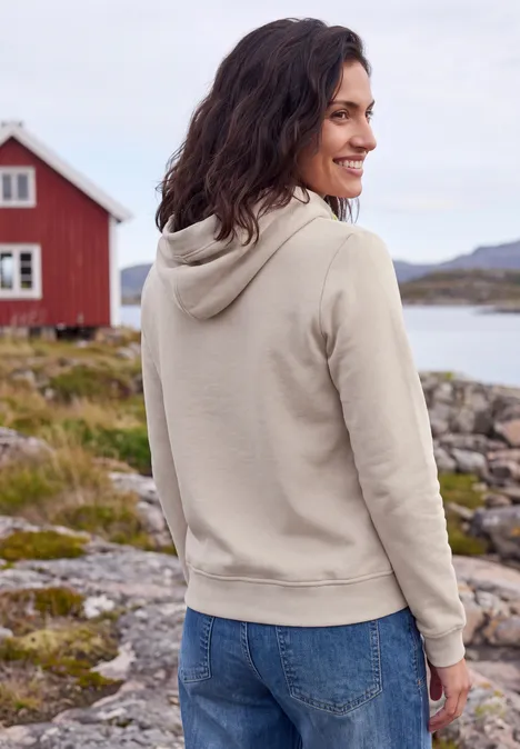 Sweatshirt mit Kapuze und Artwork snow cloud beige Frau in beigem Hoodie und Jeans steht auf felsiger Küste, lächelt über die Schulter vor rotem Haus.