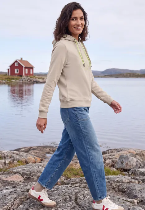 Sweatshirt mit Kapuze und Artwork snow cloud beige Eine Frau in Hoodie und Jeans spaziert am Seeufer vor einer roten Hütte.