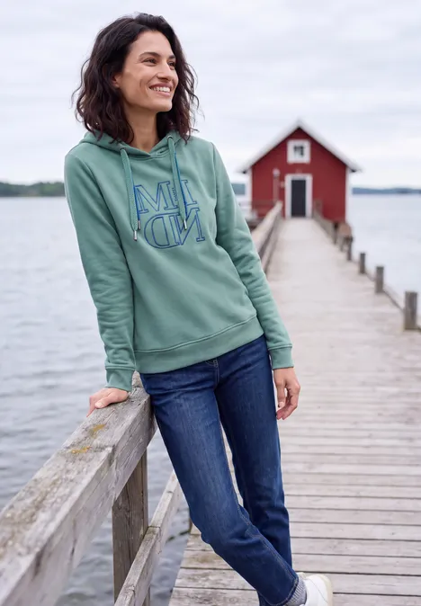 Sweatshirt mit Kapuze und Artwork mineral blue Eine Frau lehnt lächelnd an einem Steg am See, trägt grünen Hoodie und Jeans, im Hintergrund eine rote Bootshütte.