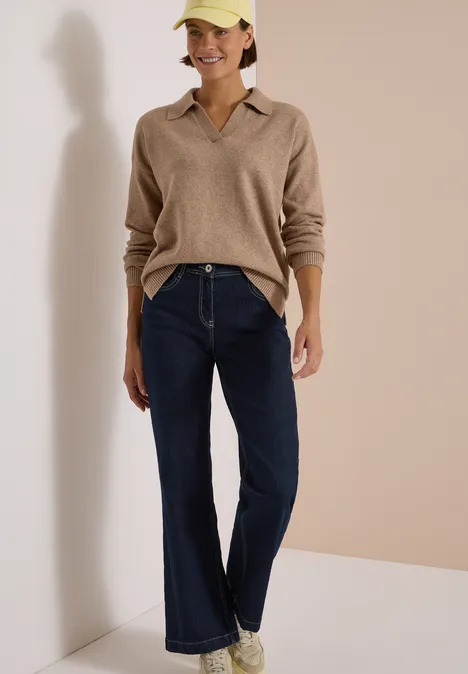 Femme en pull beige, jean large, baskets blanches et casquette jaune devant un fond neutre.