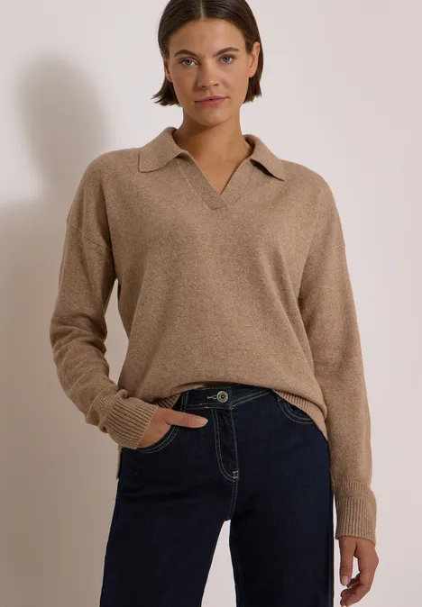 Femme portant un pull col polo beige et jean bleu foncé devant un fond clair.