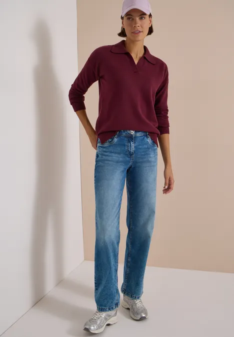 Femme en polo bordeaux, jean bleu, baskets blanches et casquette rose sur fond neutre.