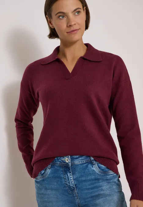 Femme portant un pull bordeaux à col en V et un jean bleu, posant devant un fond blanc.