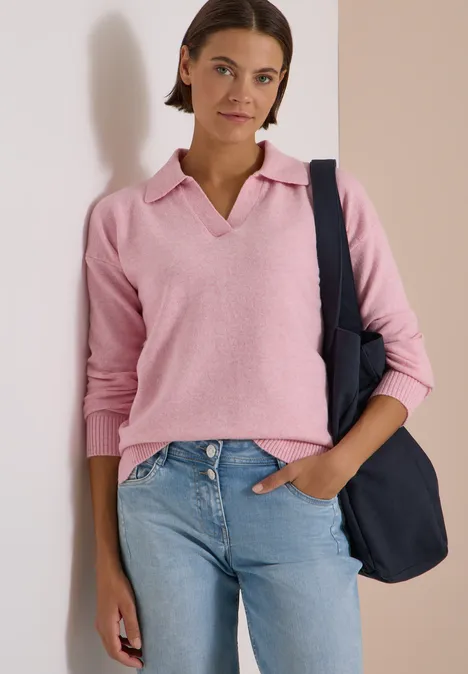 Eine junge Frau trägt ein rosafarbenes Poloshirt, Jeans und eine dunkelblaue Tasche über der Schulter.