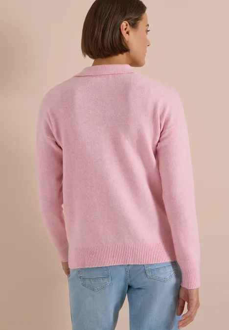 Person steht mit dem Rücken zur Kamera, trägt einen rosa Pullover und blaue Jeans vor beigem Hintergrund.