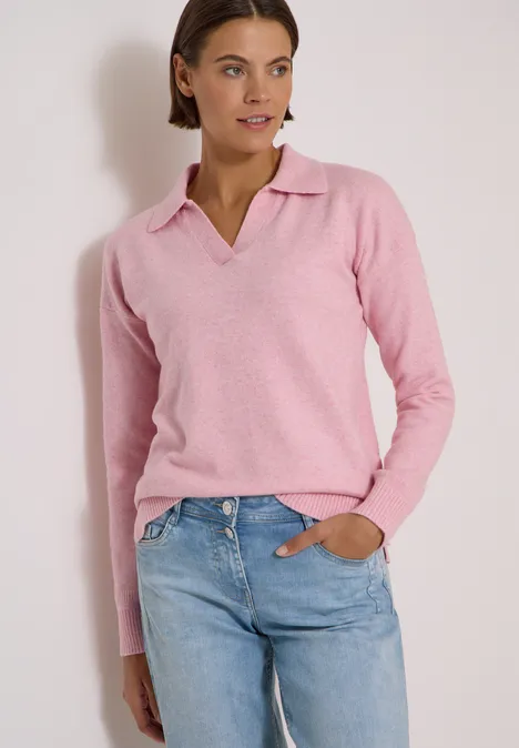 Frau trägt rosa Pullover mit Polokragen und blaue Jeans vor weißem Hintergrund.