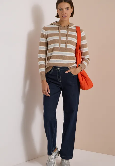 Femme en pull à capuche rayé, jean bleu et sac bandoulière rouge sur fond clair.