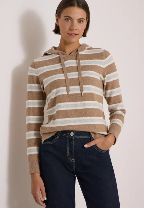 Femme en sweat à capuche rayé beige et blanc avec jean, posant devant un fond neutre.