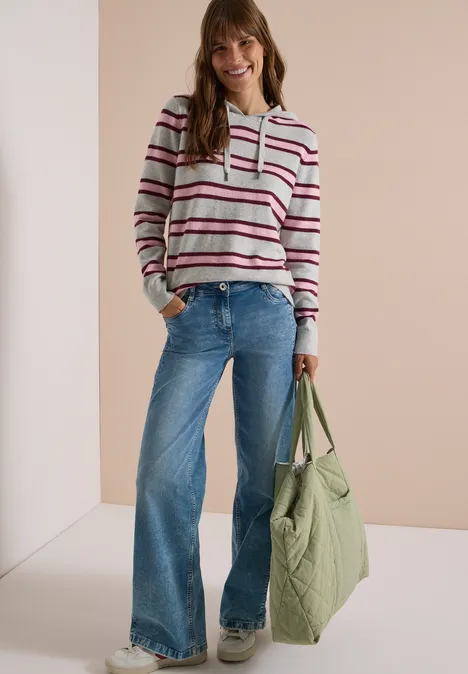 Frau mit gestreiftem Hoodie, weiten Jeans und großer grüner Tasche, lächelnd vor hellem Hintergrund.