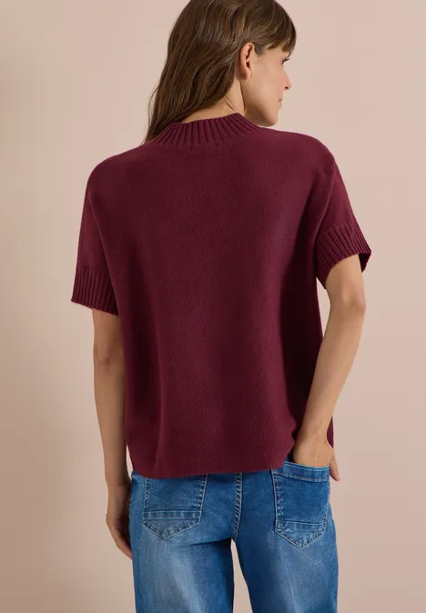 Rückansicht einer Frau in weinrotem Kurzarm-Strickpullover und Jeans vor beigem Hintergrund.