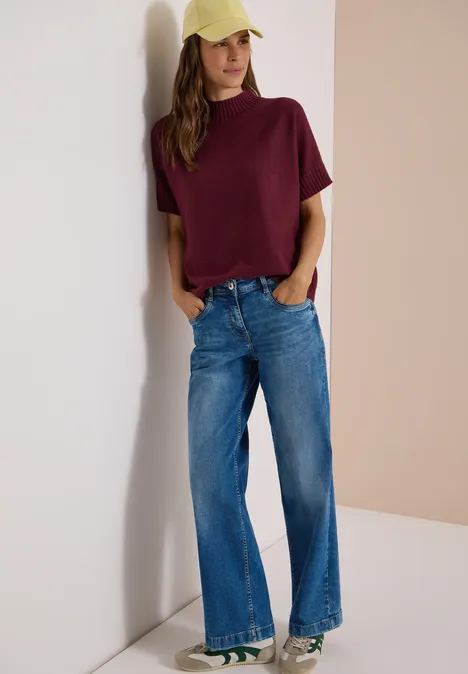 Frau lehnt an einer Wand, trägt ein bordeauxfarbenes T-Shirt, weite Jeans und eine gelbe Kappe.