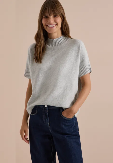 Eine lächelnde Frau in grauem Kurzarm-Strickpullover und blauen Jeans.