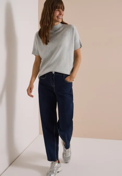Frau lehnt lässig an Wand und trägt graues T-Shirt, blaue Jeans und weiße Sneaker.