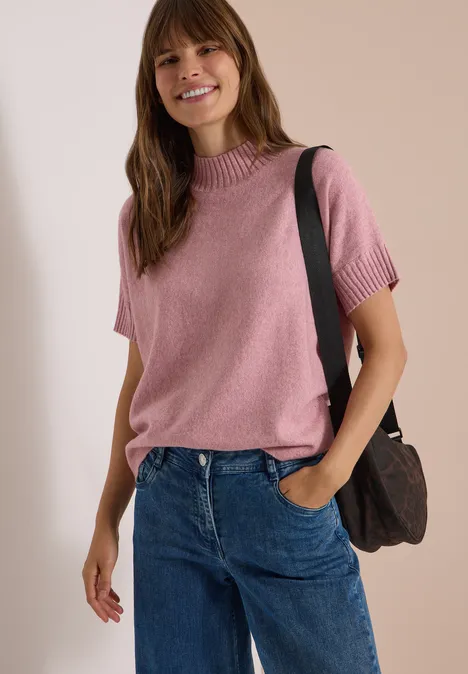 Frau in rosa Kurzarm-Pullover, Jeans und Schultertasche steht lächelnd vor neutralem Hintergrund.