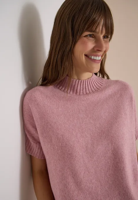 Eine lächelnde Frau lehnt an einer Wand und trägt einen rosa Kurzarm-Pullover.