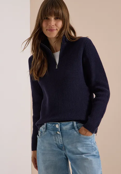 Femme vêtue d’un pull bleu marine à demi-fermeture éclair et d’un jean, posant devant un fond neutre.
