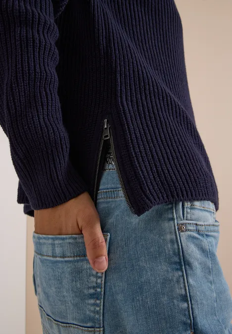 Gros plan d’un buste en pull côtelé bleu marine avec zip latéral et jean clair.