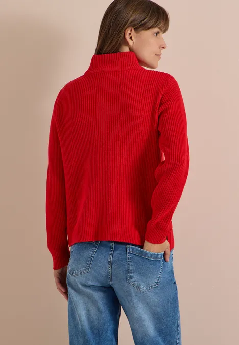 Femme de dos, regard latéral droit, pull col roulé rouge et jean bleu sur fond beige.