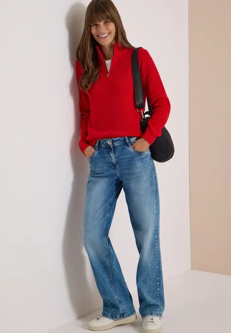Une femme en pull rouge, jean bleu et baskets, souriante, appuyée contre un mur avec un sac épaule.