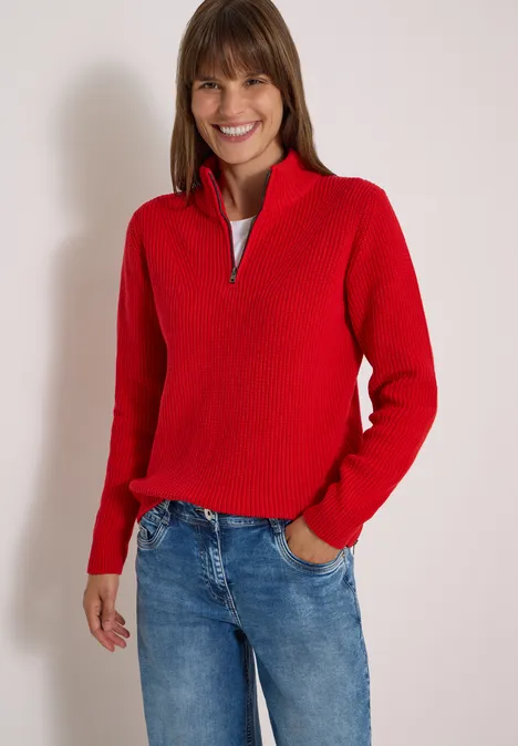 Une femme souriante porte un pull rouge et un jean bleu sur fond blanc.