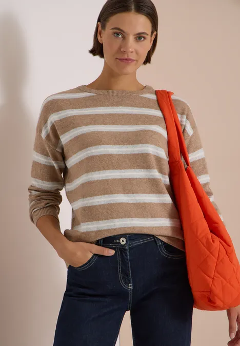 Femme en pull beige à rayures blanches, jean foncé et sac orange, posant devant un fond lumineux.