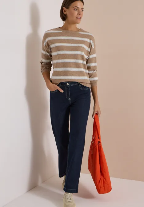 Une femme debout devant un fond neutre, pull rayé, jean large et sac orange à la main.