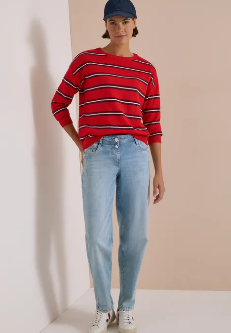 Frau mit rotem, gestreiftem Shirt, hellen Jeans, weißen Sneakern und blauer Cap vor hellem Hintergrund.
