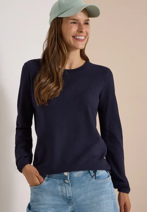 Une femme souriante porte un pull bleu marine, un jean décontracté et une casquette vert clair.