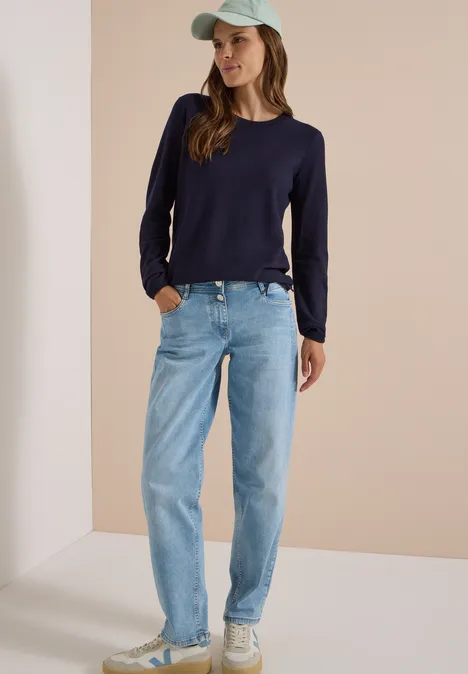 Femme en pull bleu foncé, jean clair et casquette, posant devant un mur beige clair.