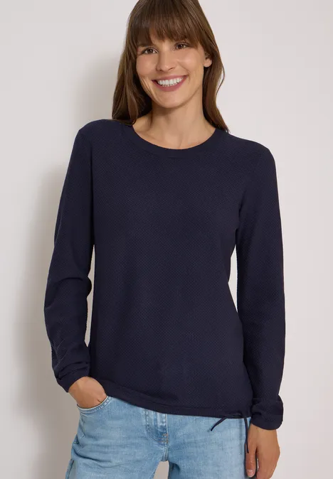Femme sourit en pull bleu marine et jean clair sur fond blanc.