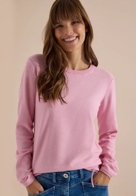 Femme souriante en haut rose à manches longues devant un fond clair.