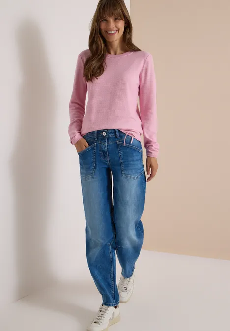 Femme portant un pull rose, un jean bleu et des baskets blanches sur fond neutre.
