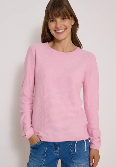 Une femme souriante portant un pull rose et un jean, mains dans les poches sur fond neutre.