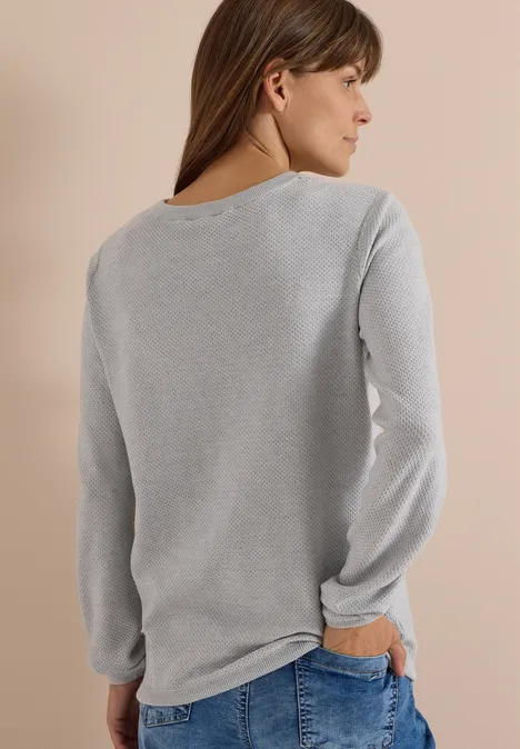 Femme de dos portant un haut gris à manches longues et un jean devant un mur beige.