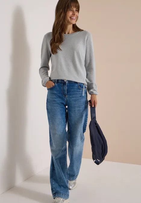 Femme souriante en haut gris, jean large et baskets blanches, tenant un sac bleu marine.