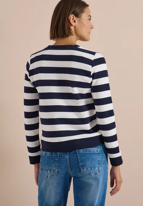 Frau von hinten in weiß-blau gestreiftem Pullover mit langen Ärmeln und Jeans vor beigem Hintergrund.