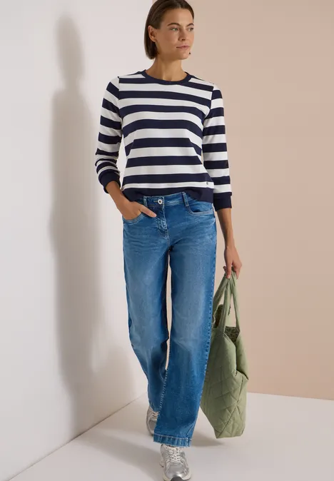 Frau trägt gestreiften Pullover, weite Jeans und hält eine grüne, gesteppte Stofftasche vor neutralem Hintergrund.
